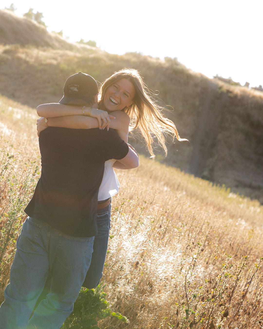 Woman laughing while hugging her partner in a sunlit grassy hillside